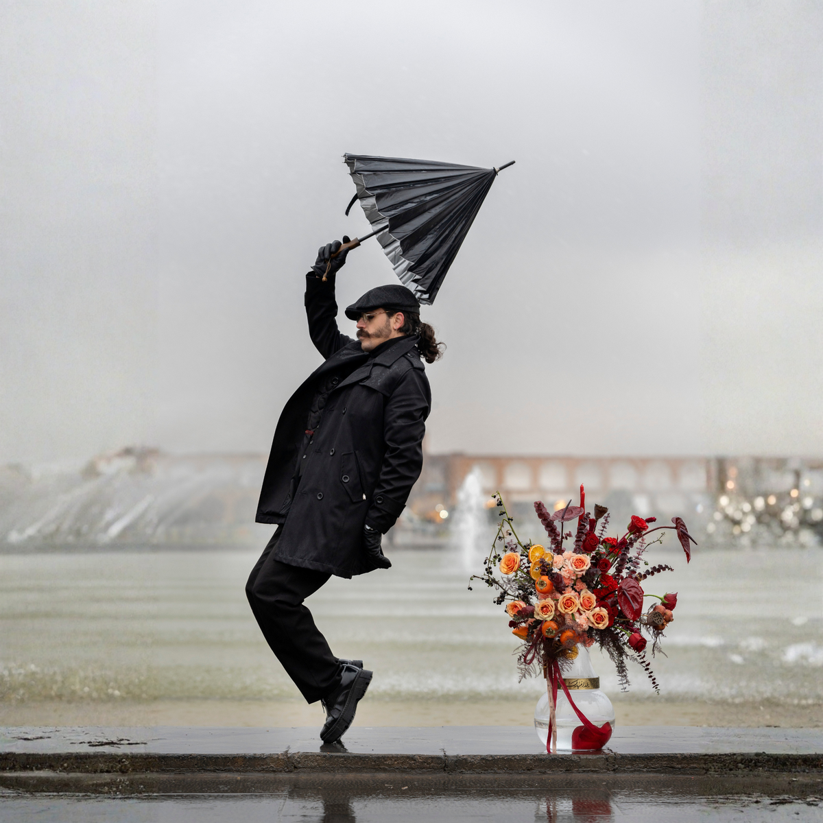 Person in black coat and hat holding a black umbrella over a bouquet of flowers on a snowy day.